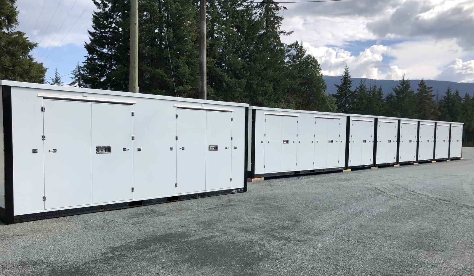 A long row of white lockable storage units on a gravel pad at Karlite Storage, with fir trees and mountains behind